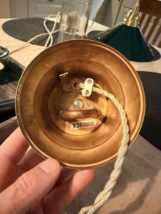 Hand håller en mässingsskärm för lampa med en textilsladd. Bakgrunden visar en grön lampskärm och en flaska på ett bord.