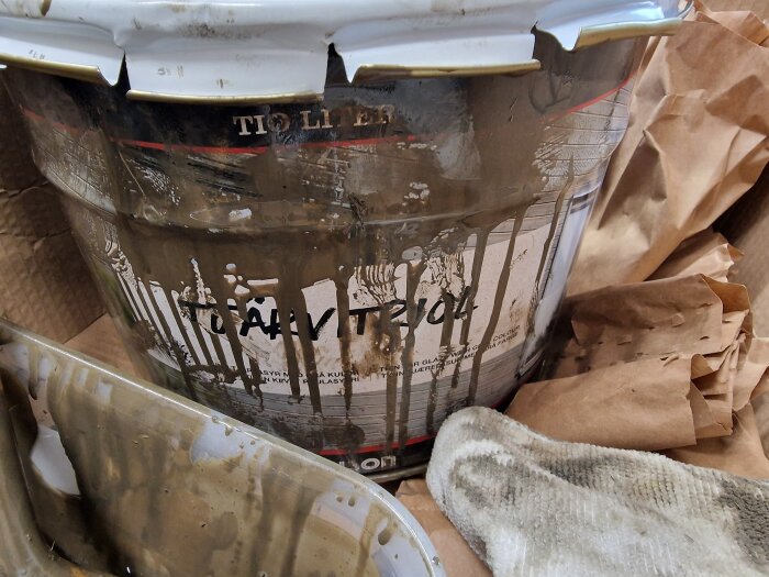 Hink med tjärvitrol, 10 liter, med färgstänk och en smutsig trasa på brun papp. Använt för utomhusmålning med svarta detaljer.