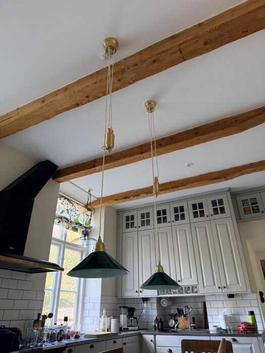 Grön kökslampa hänger från taket med träbjälkar, omgiven av vita skåp och fönster. Köksbänkar med glas och hushållsprodukter syns.