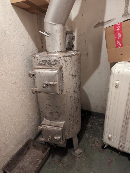 Gammal kamin i ett källarrum, ansluten till skorstenen. Ytan är nött och den står bredvid en kartong och en resväska.