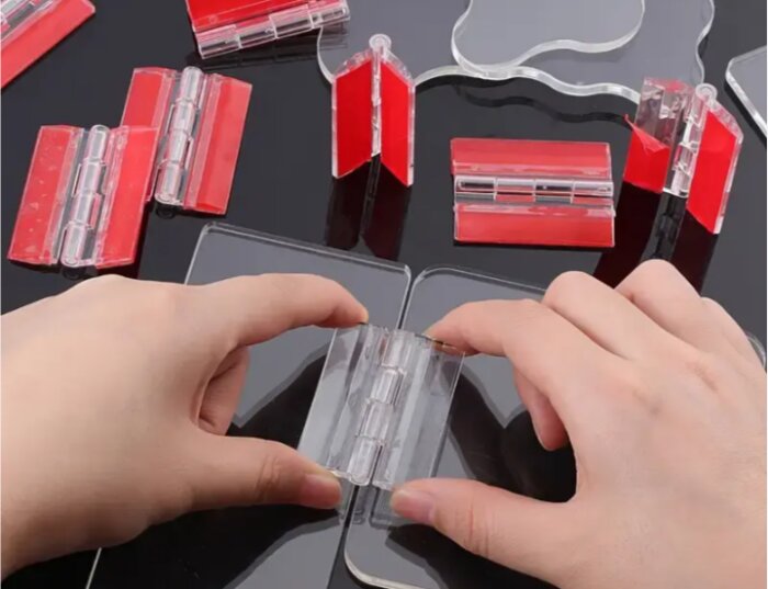 Genomskinliga akrylgångjärn med röda tejpbitar visas på ett bord, några hålls av händer.