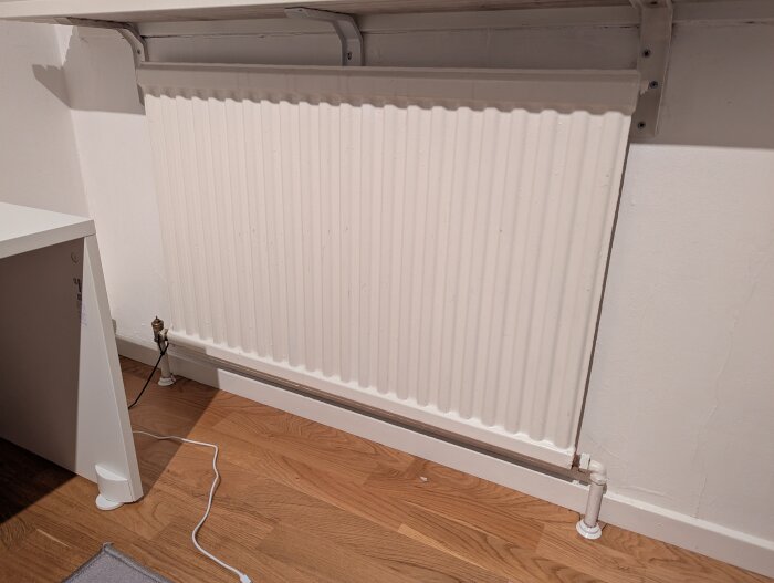 Radiator monterad mot vit vägg, rör anslutna nertill, träbeklädd golv, del av skrivbord synligt till vänster.
