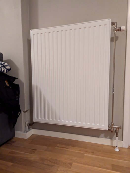 En vit väggmonterad radiator med anslutande rör, placerad ovanför ett trägolv.