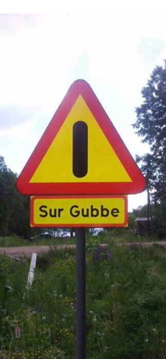 Varningsskylt med texten "Sur Gubbe" stående vid en vägkant omgiven av grönska.