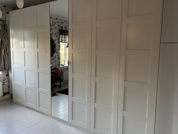 Nymonterade garderobsdörrar med spegeldörr i mitten, vita panelskåp på vardera sida, i ett sovrum med mönstrad tapet och ljus inredning.