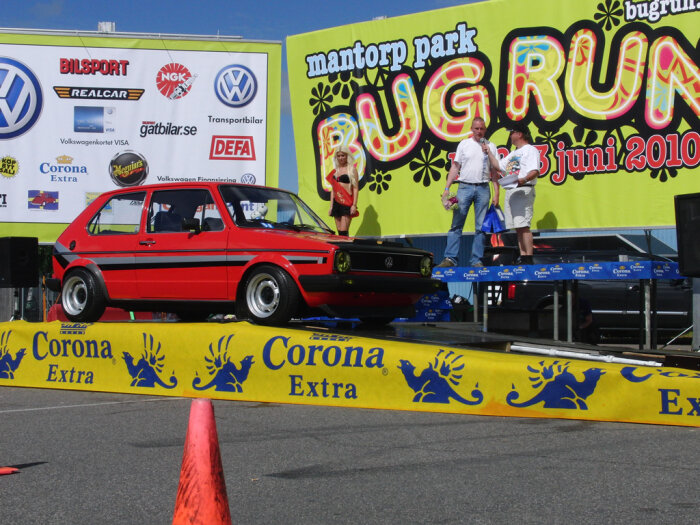 Röd bil ställs ut på Bug Run på Mantorp Park; människor ses samtala på en scen med ett färgglatt banderoll i bakgrunden.