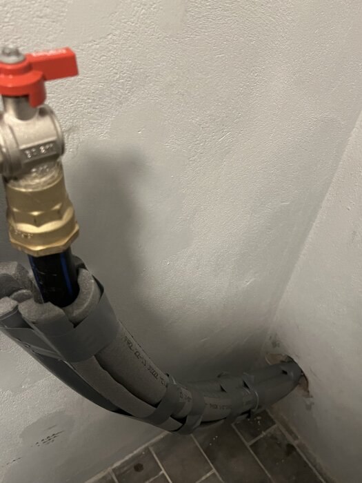 Inkommande vattenledning med rörrisolering inkapslad i grått skydd, ansluten genom ett hål i väggen, med en röd ventil.