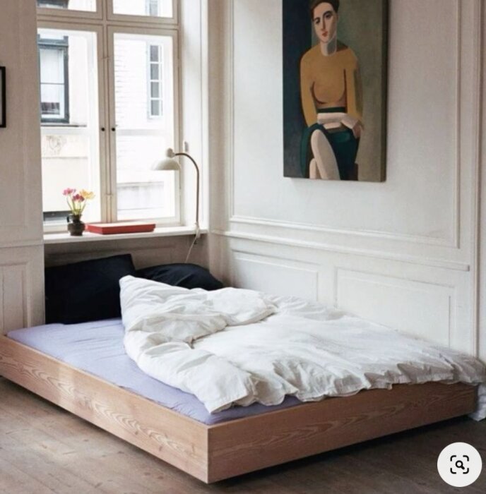 Låg träram för sängbotten, placerad direkt på golvet i ett ljust rum med vita väggar och en stor tavla ovanför.