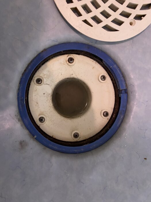 Golvbrunn med skruvar i ett badrumsgolv täckt med plastmatta; vattenlåset synligt utan handtag eller skarvar.