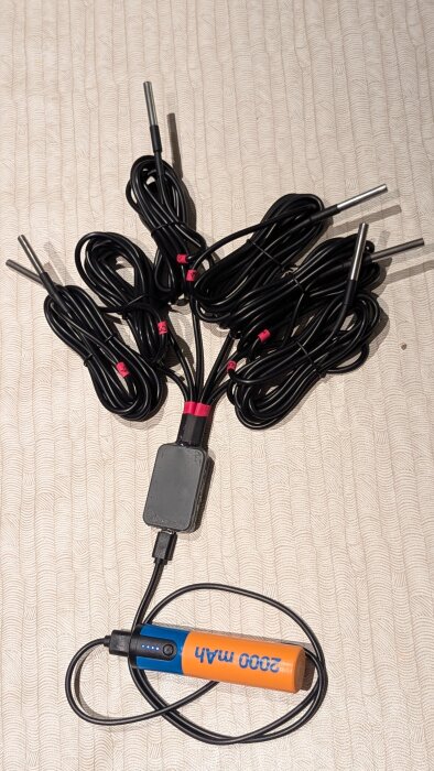Flera svarta sensorkablar med stålprober och en batteribank på 2000 mAh ligger utspridda på ett mönstrat tygunderlag.