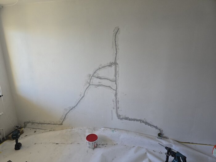 Vägg med torkande bruk och borr förberedd för målningsarbete. Golvet täckt med skyddsplast och en rödvit hink synlig.