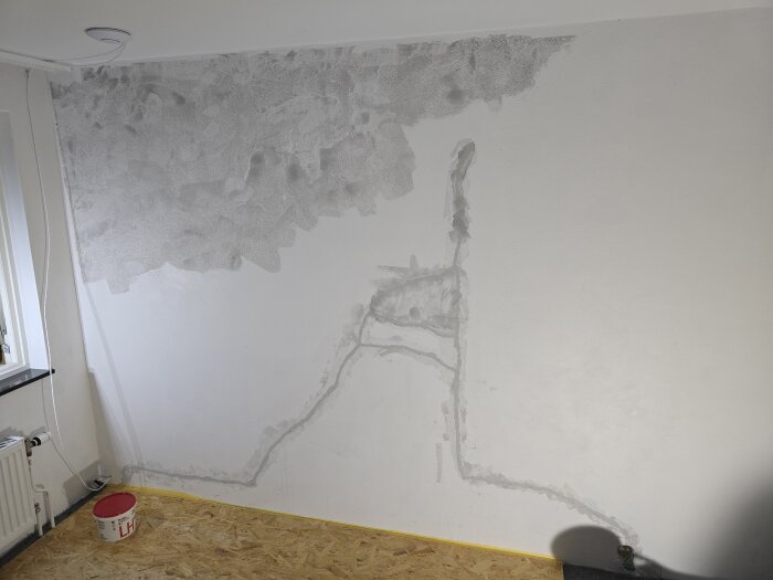 Vägg med torkande spackel och eldragning, vita kablar längs väggen, hink på golvet, fönster till vänster.