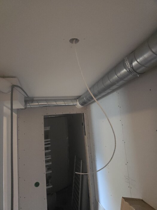 Ventilationsrör vid tak med hängande kabel för takbelysning i ett rum under renovering, synliga väggar och öppning till annan del av huset.