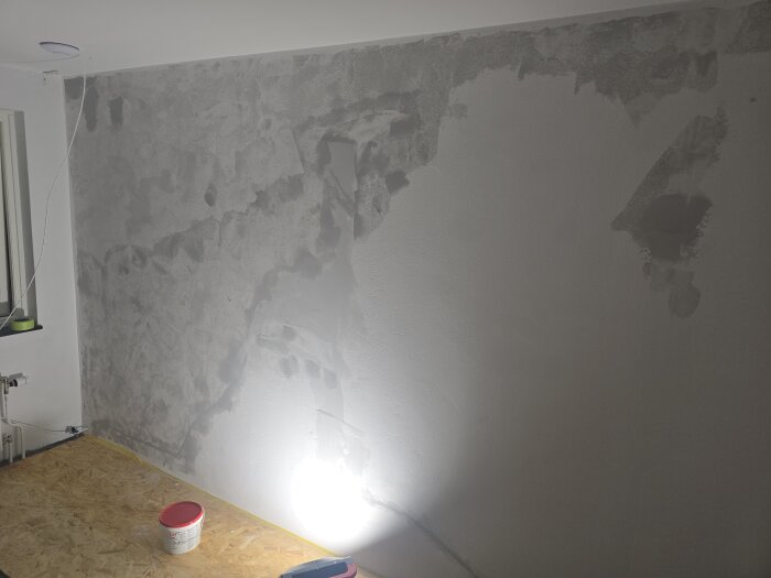 Vägg under bredspackling med ojämnheter och belysning på golvet. En burk med rött lock står på en träskiva.