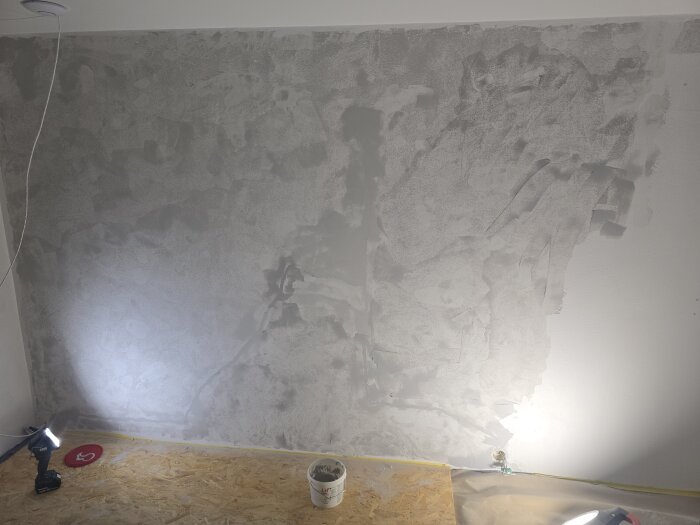 Vägg under spacklingsprocess, med elektrisk verktyg och spackelhink på golvet. Belysning avslöjar torkande yta och strukturer.
