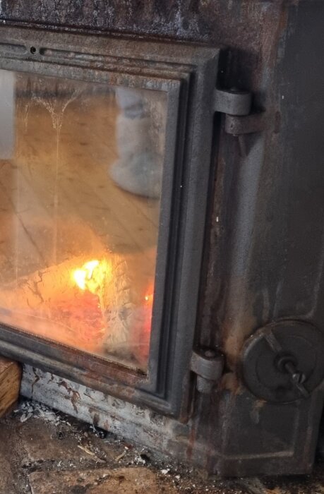 Gångjärnet på en eldstadsdörr har gått av, synlig låga innanför glaset.
