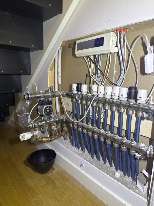 Cirkulationspump med rör och ventiler under trappa, svart skål på golvet nedanför, elcentral i bakgrunden.