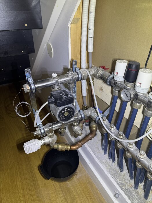 Cirkulationspump med anslutna rör och ventiler i ett hem, placerad ovanför en svart hink, mot en vägg med blåa rör i bakgrund.