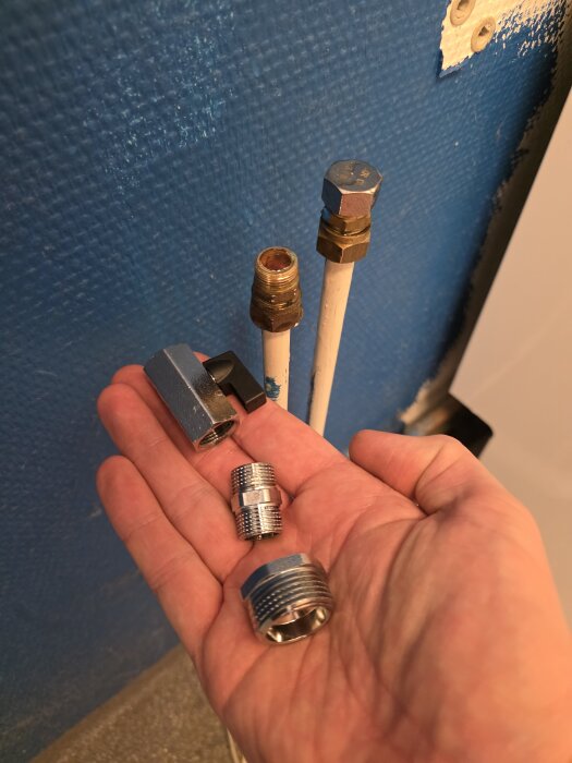 Hand som håller i tre olika rörkopplingar framför två rör monterade på en blå vägg; en koppling med svart komponent syns.