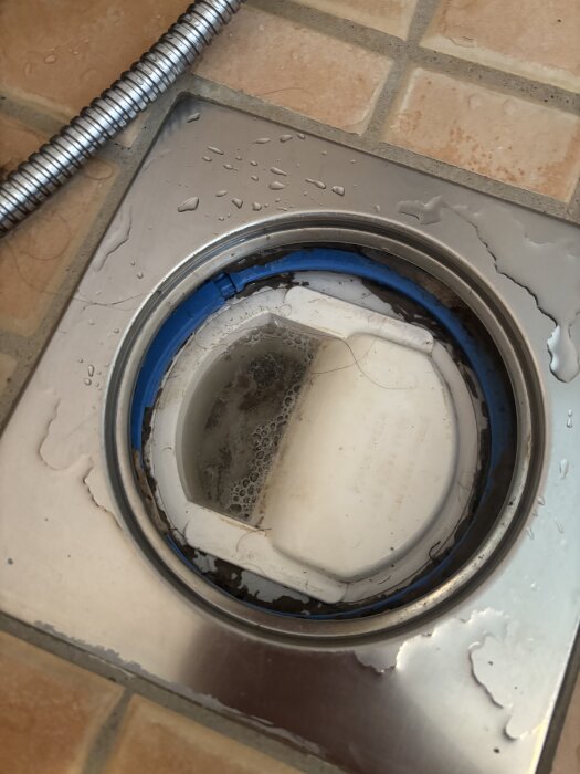 Golvbrunn i badrum från 2002 med vatten och smuts, omgiven av kakel och metallram, och en blå tätning som verkar trasig.