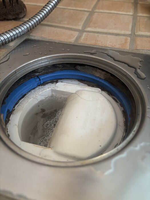 Golvbrunn med synlig blå ring och små mängder vatten med smuts och hår, i ett badrum från 2002 med klinkergolv.