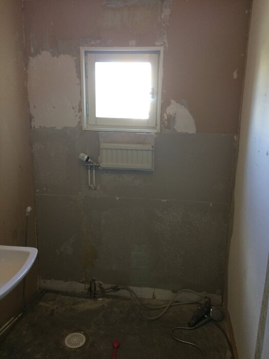 Orenoverat badrum med delvis rivna väggar och gamla rör, fönster ovanför en radiator, duschgolv med golvbrunn och lös duschslang på golvet.