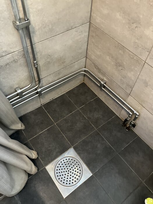 Rörinstallationer längs vägg och golv i ett badrum med grå kakelplattor och duschavlopp synligt.