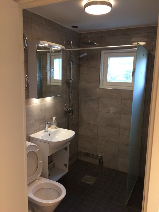 Badrum med grå kakel, dusch med glasdörr, handfat, toalett, och rörgenomföring nära golvet.