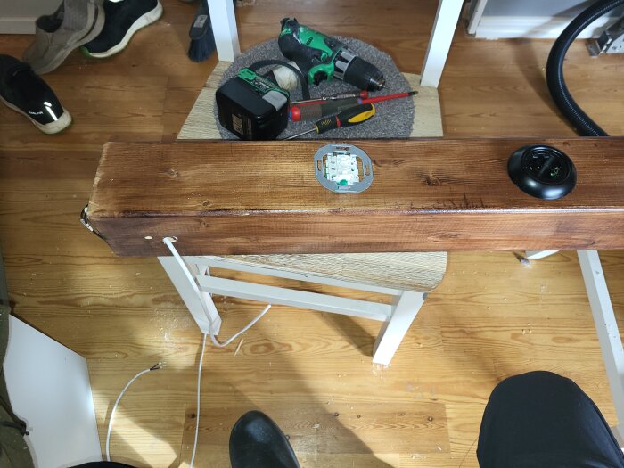 Träplanka med pågående elinstallation, synlig kabel och verktyg i bakgrunden på ett trägolv, omgiven av diverse föremål.