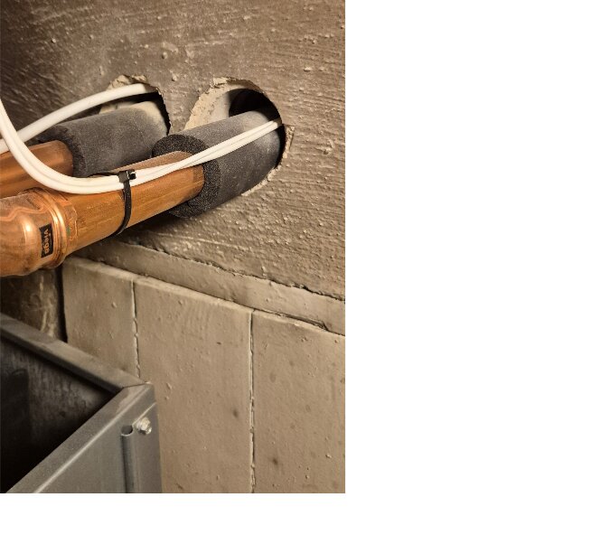 Genomföringar för rör och kablar genom en vägg, kopplade till en luft/vatten-värmepump, med isoleringsmaterial runt rören.