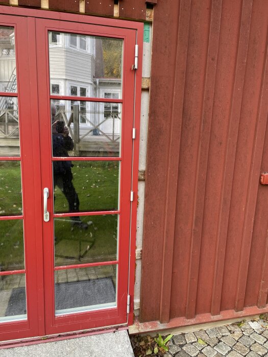 Röd glasad pardörr från Ekstrands med justeringsproblem i gångjärn, monterad i en träfasad. Reflexion av fotograf och vitt hus i bakgrunden.
