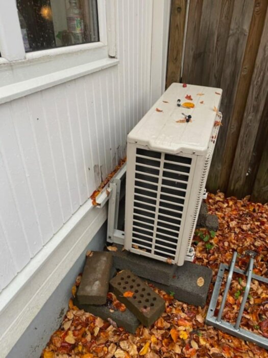 Luftvärmepumpens utomhusaggregat som fallit till marken, vilande på lecablock, mot husfasad med höstlöv omkring.