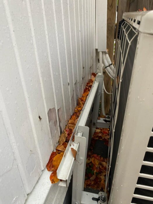 Luftvärmepumps utomhusdel som har lossnat och står på marken, omgiven av höstlöv vid en vägg.