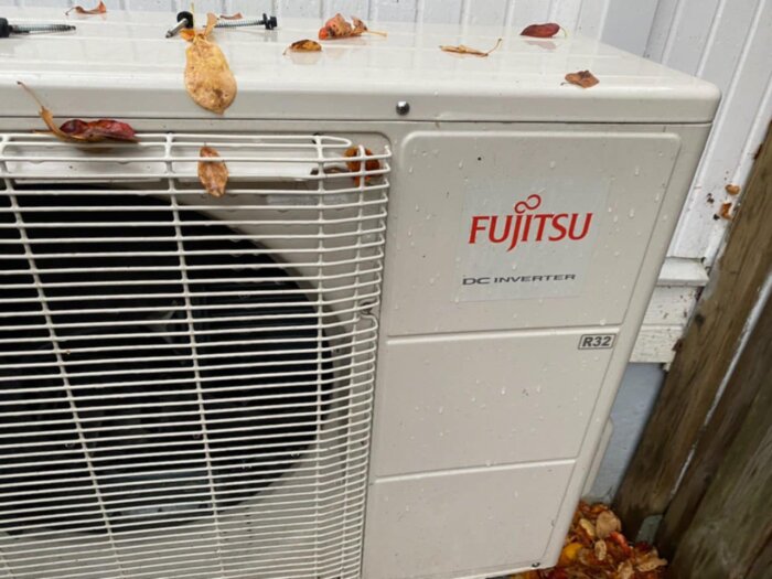 Luftvärmepump med Fujitsu-logga placerad på marken, omgiven av löv och vägg i bakgrunden.