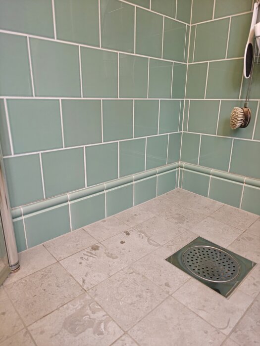 Grönt kakel i dusch med sockel och beige golvklinker, duschbrunn synlig.