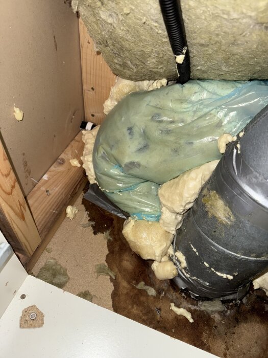 Oisolerat metallrör med kondens och vattenskada bakom borttagen byrå, synligt med träreglar, isolering, plaströr och fuktskadad yta.