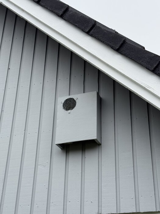 Ventilationsutlopp med galler monterat på yttervägg av en byggnad med liggande träpanel och svart tak.