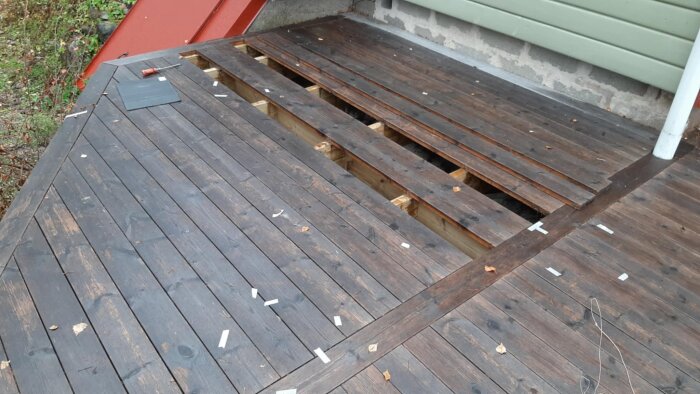 Öppet trädäck med synliga bjälkar där några plankor saknas, omgiven av markeringar och verktyg.