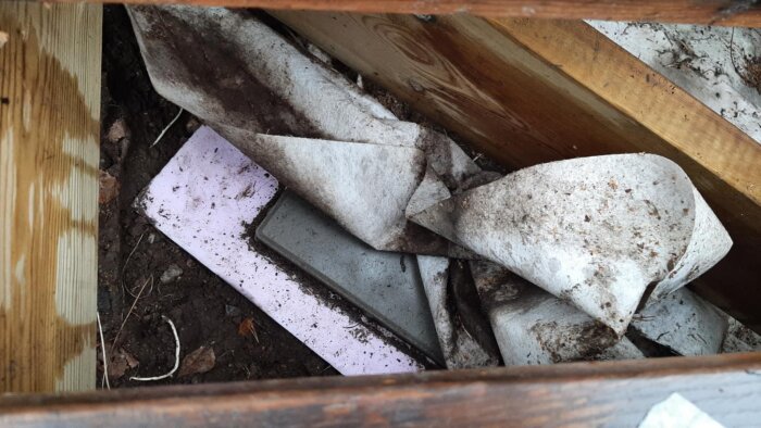 Byggmaterial under trädäck med jord, cellplastskivor och betongplattor.