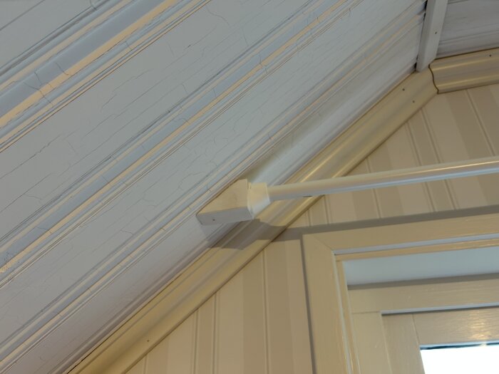 Gardinstång monterad i snedtak vid ett fönster med ljusa träpaneler. Enkel lösning för att få gardiner att hänga längre ut.