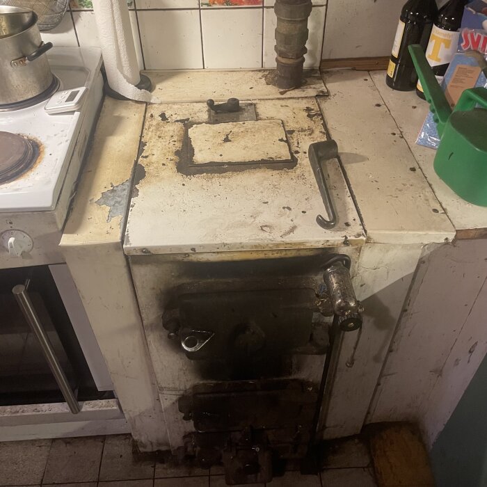 Gammal vedspis med rostiga detaljer och bränd yta, omgivet av en kastrull, en ugn och några flaskor i ett kök.