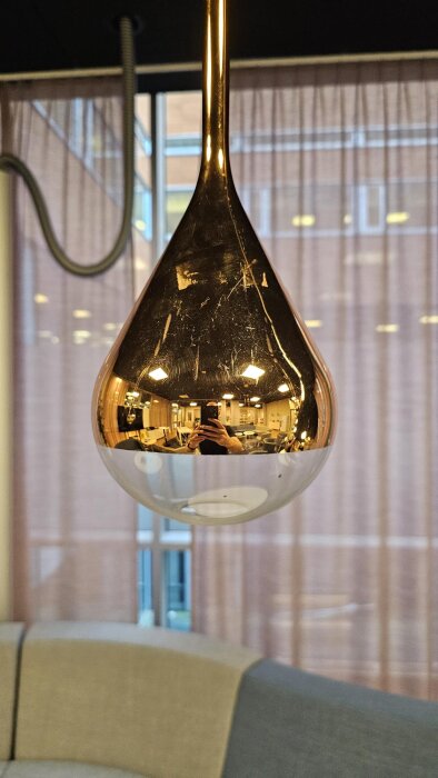 Glödlampa i kopparfärgad armatur med reflekterande yta, synlig i kontorsmiljö med soffa och fönster i bakgrunden.