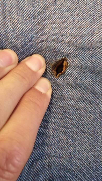 Hand vid bränt hål i tygklädsel, nära synlig metall.
