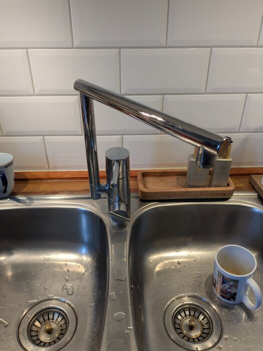 Kökskran i stål ovanför dubbel diskho, med vattenstänk och en mugg i en av hoarna.