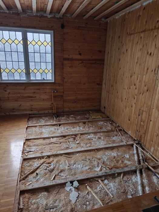 Rum under renovering med blottat undergolv och träpanelade väggar, samt ett fönster med färgat glas.