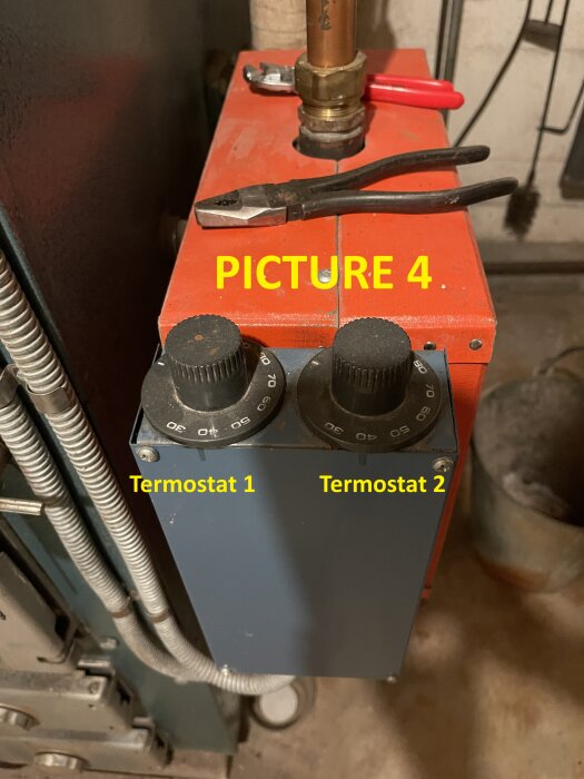 Elektrisk värmare med två termostater märkta "Termostat 1" och "Termostat 2", placerade ovanpå en röd enhet med en rörtång och samtidigtänger.