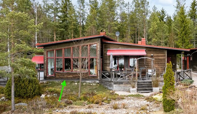 Trähus med stora fönster och röda markiser, omgiven av skog och trädgård. Snett tak över fönsterväggen.