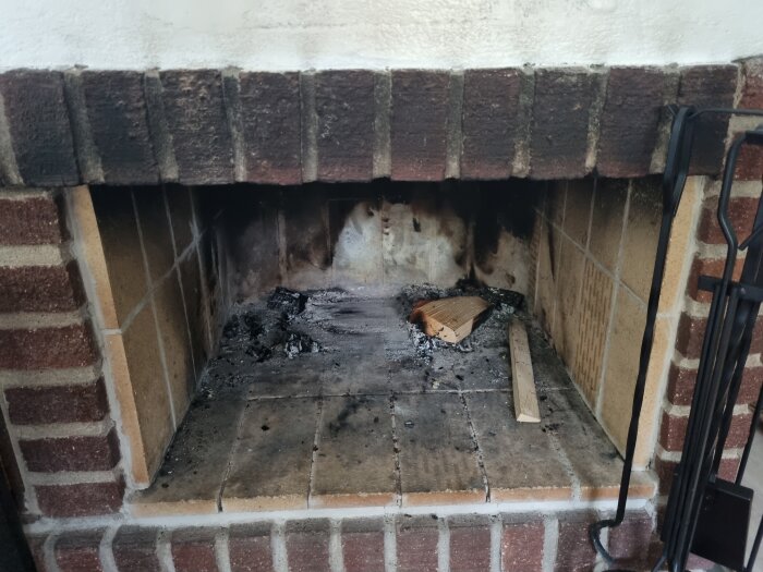 En öppen spis i tegel med sotspår, aska och en bit obehandlat trä ligger kvar på botten.