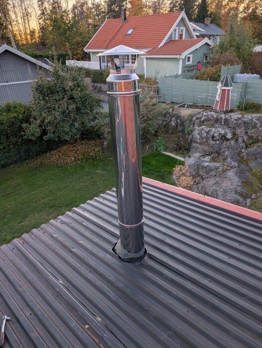 Rostfri skorsten installerad på ett plåttak, omgiven av en trädgård och flera hus i bakgrunden.