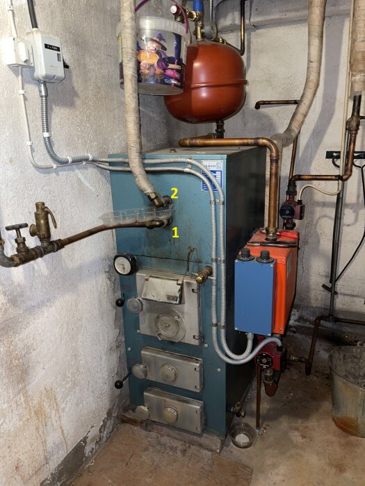 Wood boiler system with pressure gauge, pipes, and valve in a basement setting. A red expansion vessel is visible above the boiler.